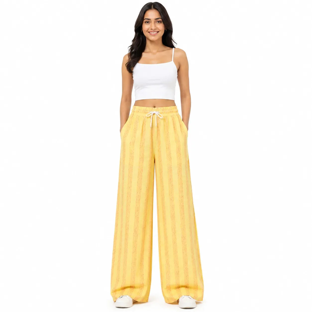 A woman modeling a pair of wide-leg yellow pants with vertical stripes and a white drawstring, paired with a white crop top and white sneakers, against a plain white background.