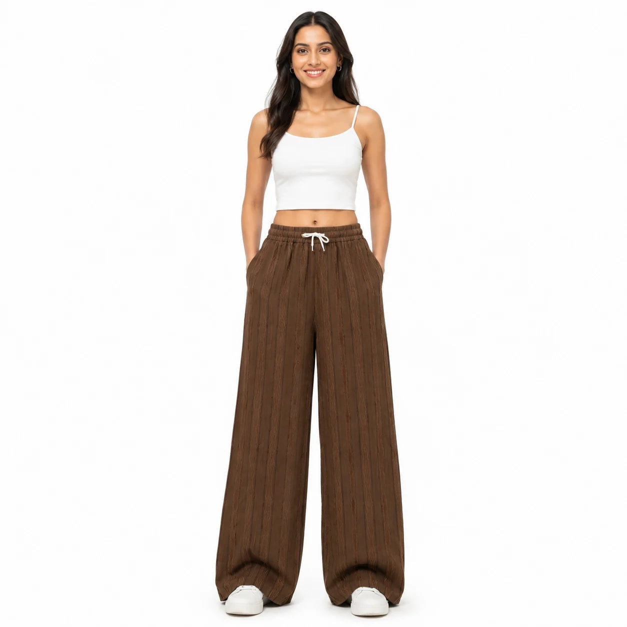 A woman modeling a pair of wide-leg brown pleated pants with a white drawstring, paired with a white crop top and white sneakers, against a solid white background.