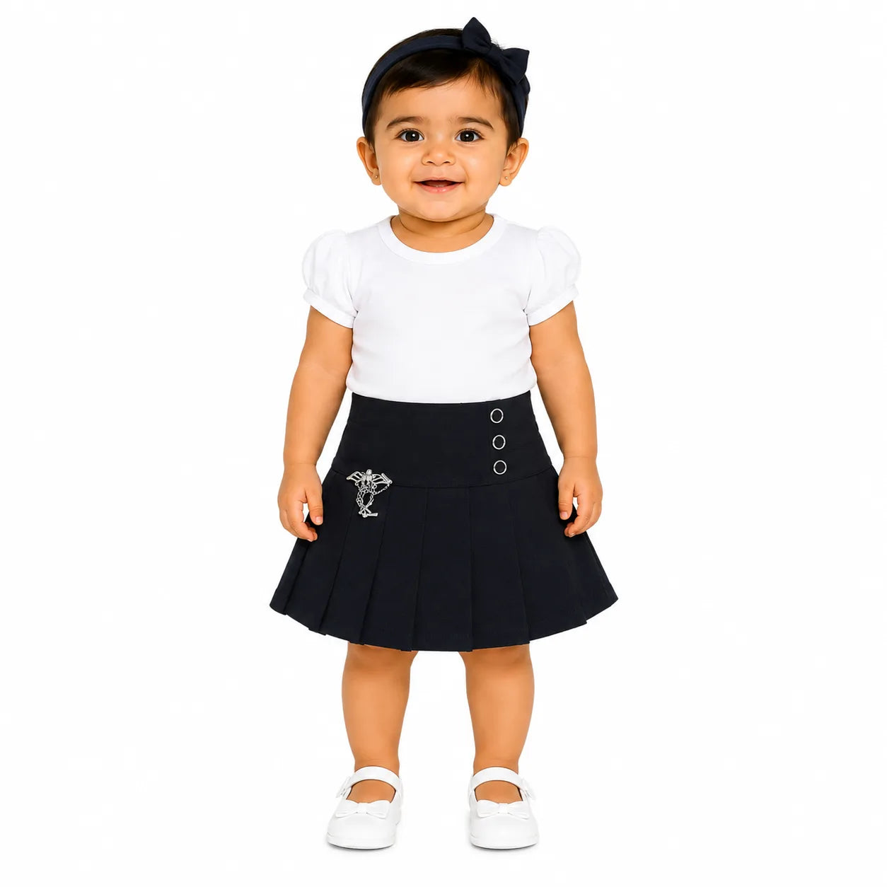 A toddler girl wearing a white short-sleeve t-shirt, a black pleated skirt with a silver embroidered crest and button details, white Mary Jane shoes, and a black bow headband, standing against a solid white background.