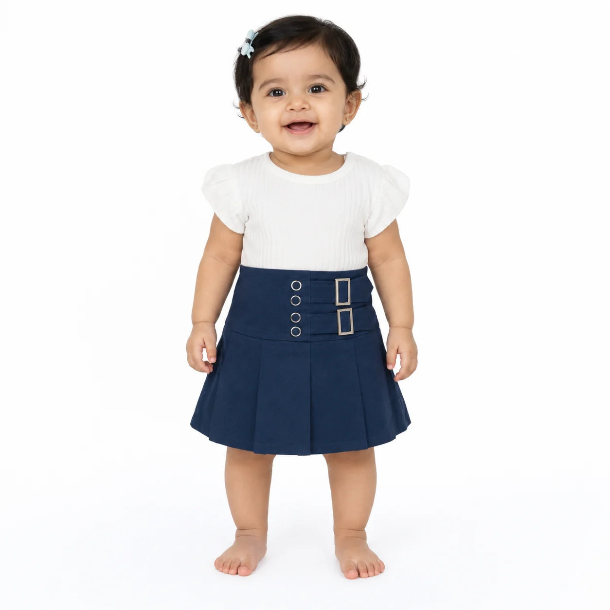 A toddler girl standing barefoot against a white studio background, wearing a white short-sleeve top and a navy pleated skirt with a decorative waistband featuring gold-tone buttons and a rectangular buckle.
