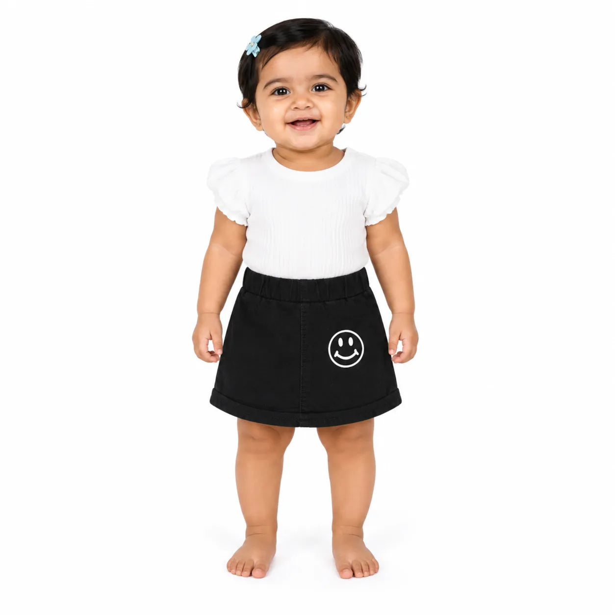 A toddler girl standing against a white background, wearing a white short-sleeve top with puffed shoulders and a black mini skirt featuring a white smiley face graphic on the left side.