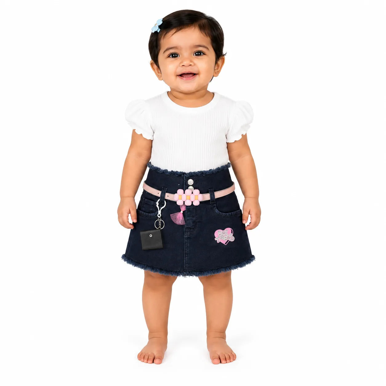A toddler girl standing against a white studio background, wearing a white short-sleeve ribbed top and a dark denim skirt with a pink floral belt, pink tassel, and a pink butterfly patch.