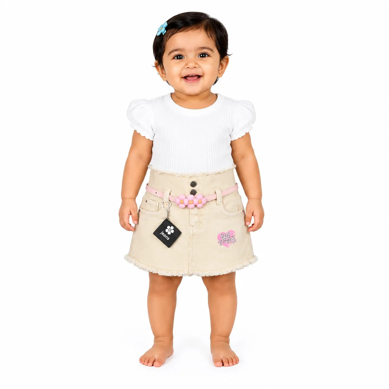 A toddler girl standing against a white background, wearing a white short-sleeve top and a beige denim skirt with a pink flower belt, a black paw-print patch, and a pink embroidered patch.