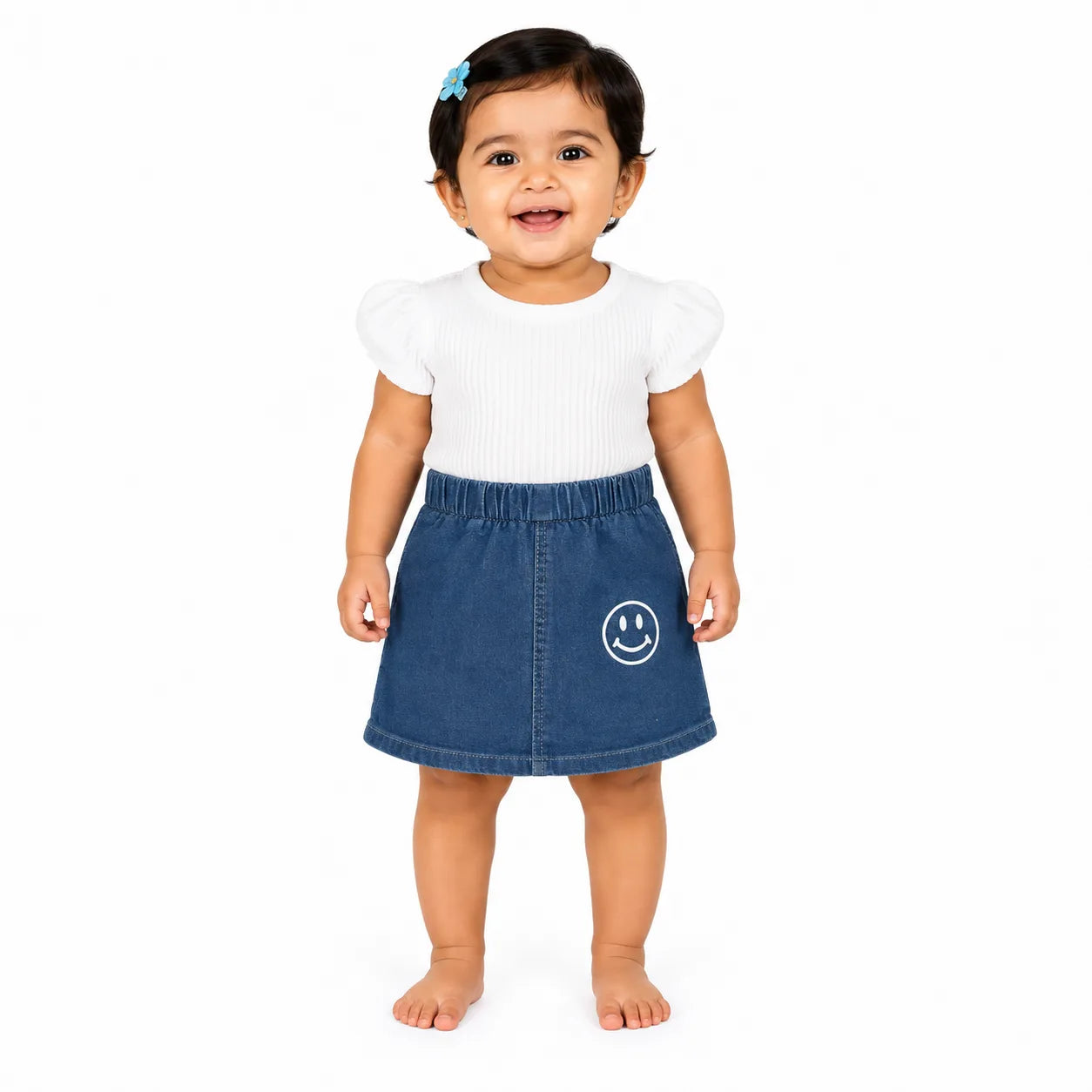 A toddler girl standing against a white background, wearing a white short-sleeve ribbed top with puffed shoulders and a blue denim skirt featuring a white smiley face graphic on the front.