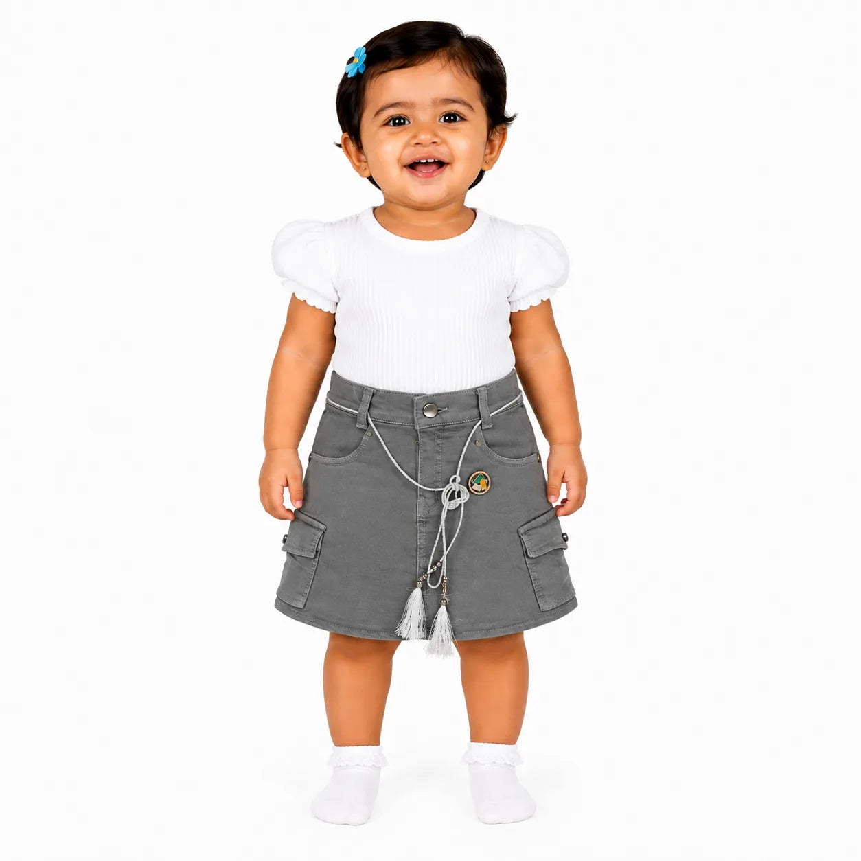 A toddler girl standing against a white studio background, wearing a white short-sleeve ribbed top with puffed shoulders and a gray cargo-style skirt with a decorative chain and tassel detail.
