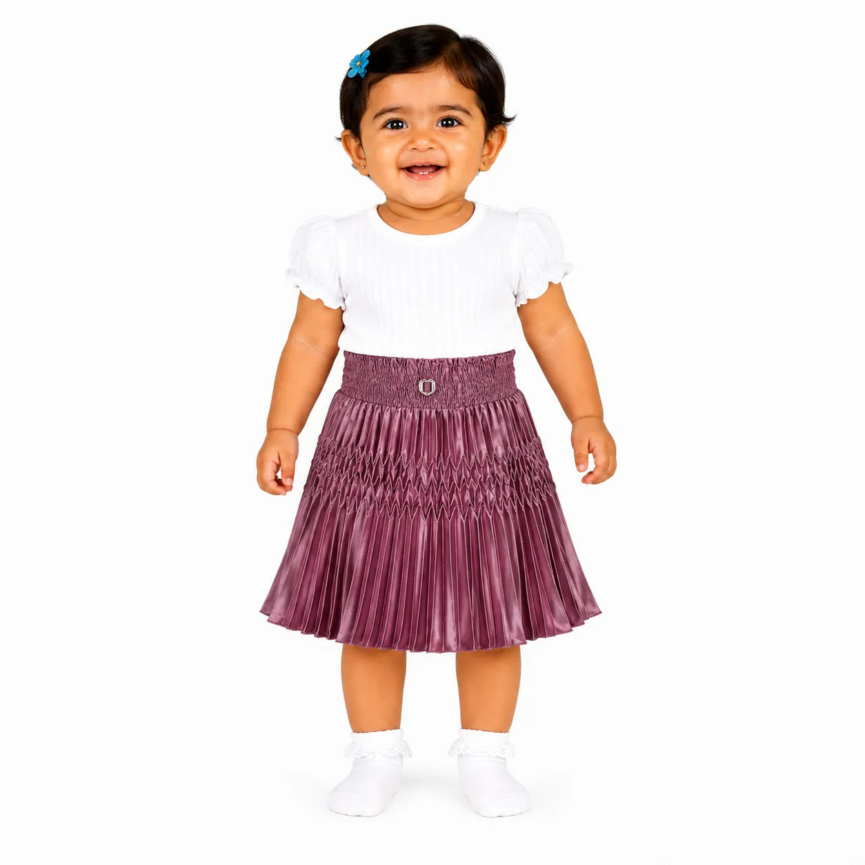 A toddler girl wearing a white short-sleeve top and a pleated metallic mauve-purple skirt, standing against a plain white background.