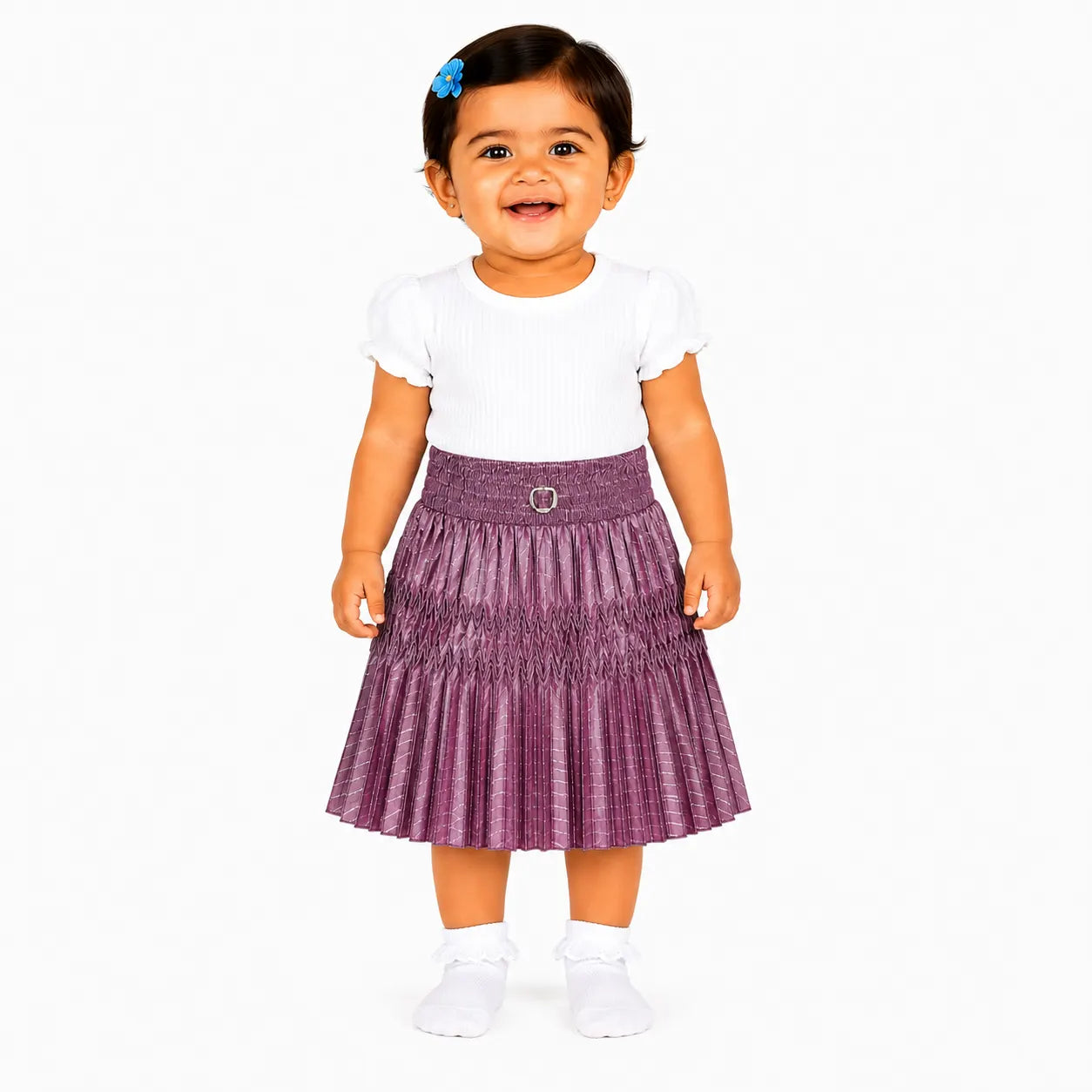 A toddler girl smiling and posing in a white short-sleeve top and a pleated purple skirt with a small silver buckle at the waist, set against a plain white background.