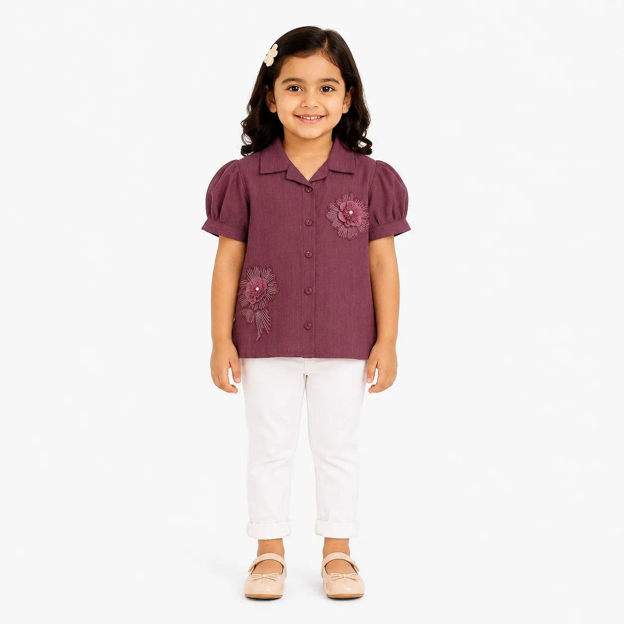 A young girl with dark hair wearing a short-sleeve button-up blouse in a dusty plum purple color with floral embroidery, paired with white pants and beige flats, standing against a plain white background.
