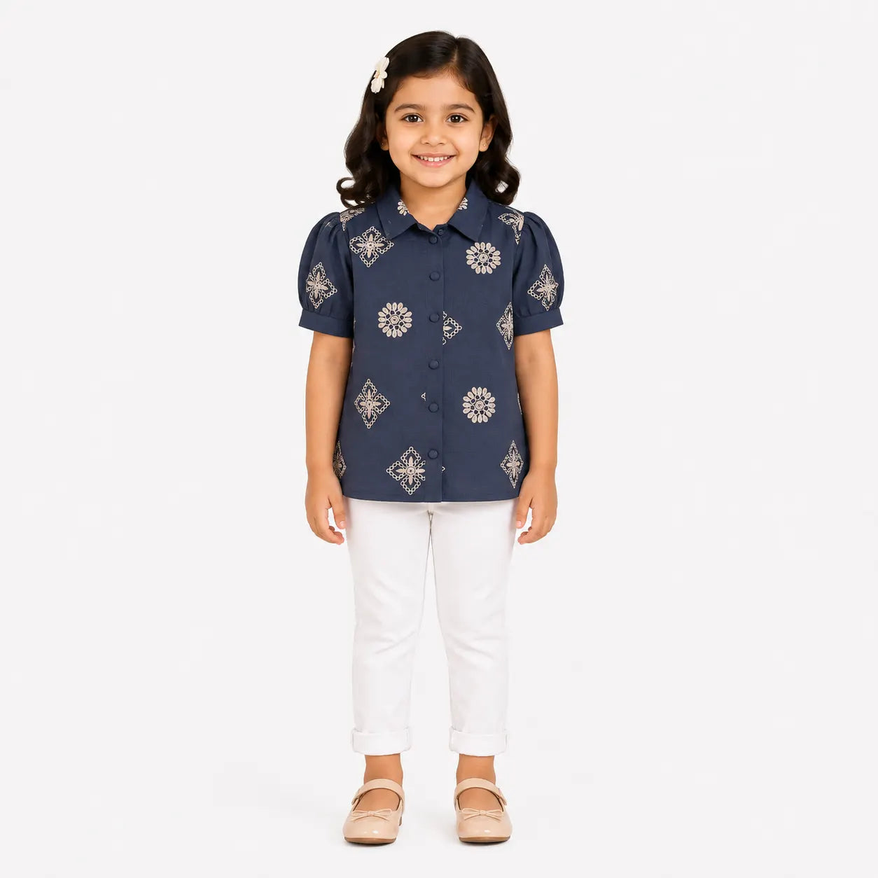 A young girl stands against a light gray studio background wearing a navy short-sleeve button-up shirt with beige geometric and floral patterns, white pants, and beige flats.