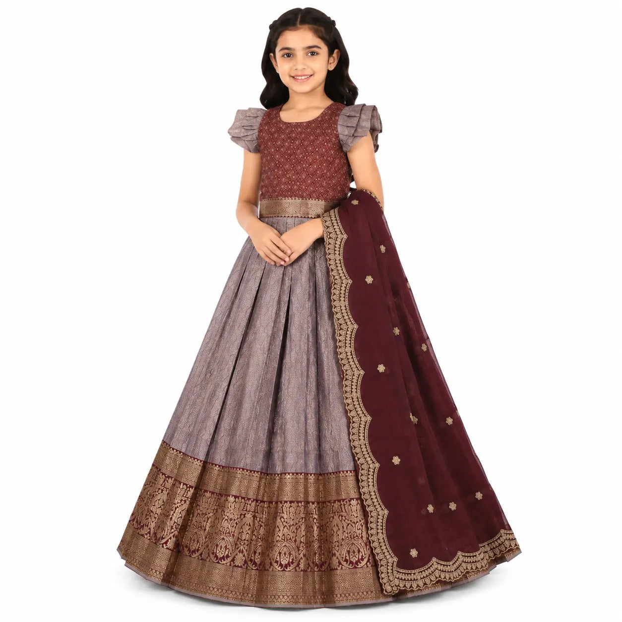 A young girl modeling a traditional Indian ethnic outfit consisting of a dusty purple-gray flared skirt (lehenga), a matching patterned blouse with ruffled sleeves, and a deep maroon dupatta with gold embroidery.