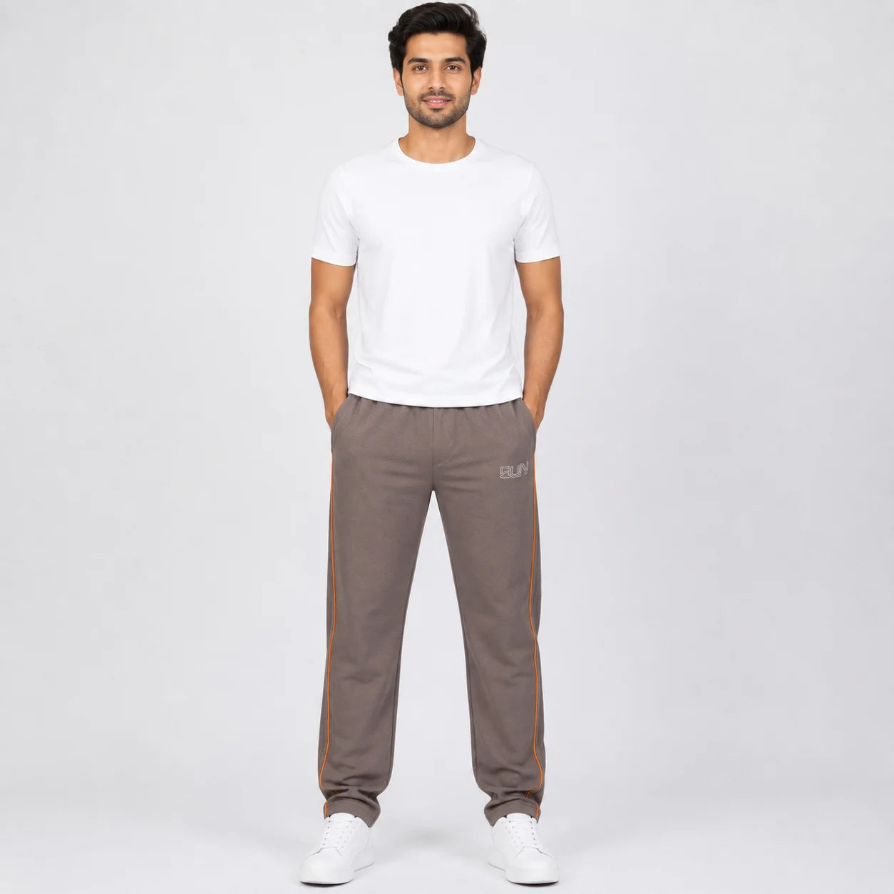 A man modeling a white crew-neck t-shirt and brown track pants with orange side stripes, standing against a light gray studio background.