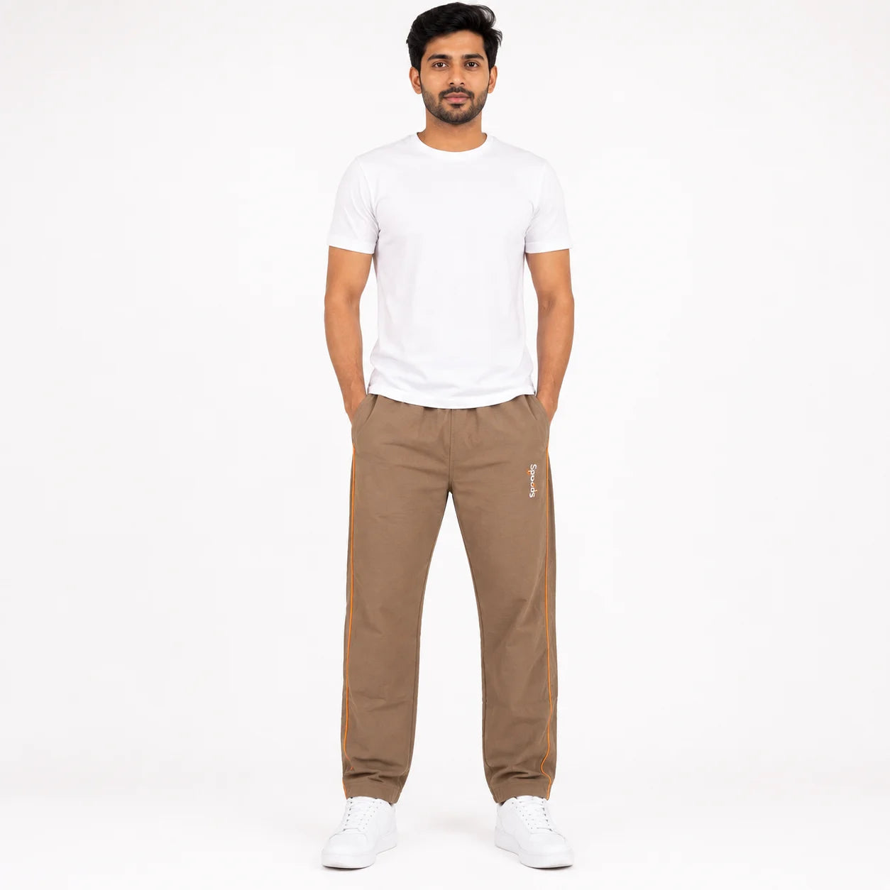 A man modeling a pair of brown jogger pants with a small white embroidered logo on the left thigh, paired with a white t-shirt and white sneakers, against a plain white background.