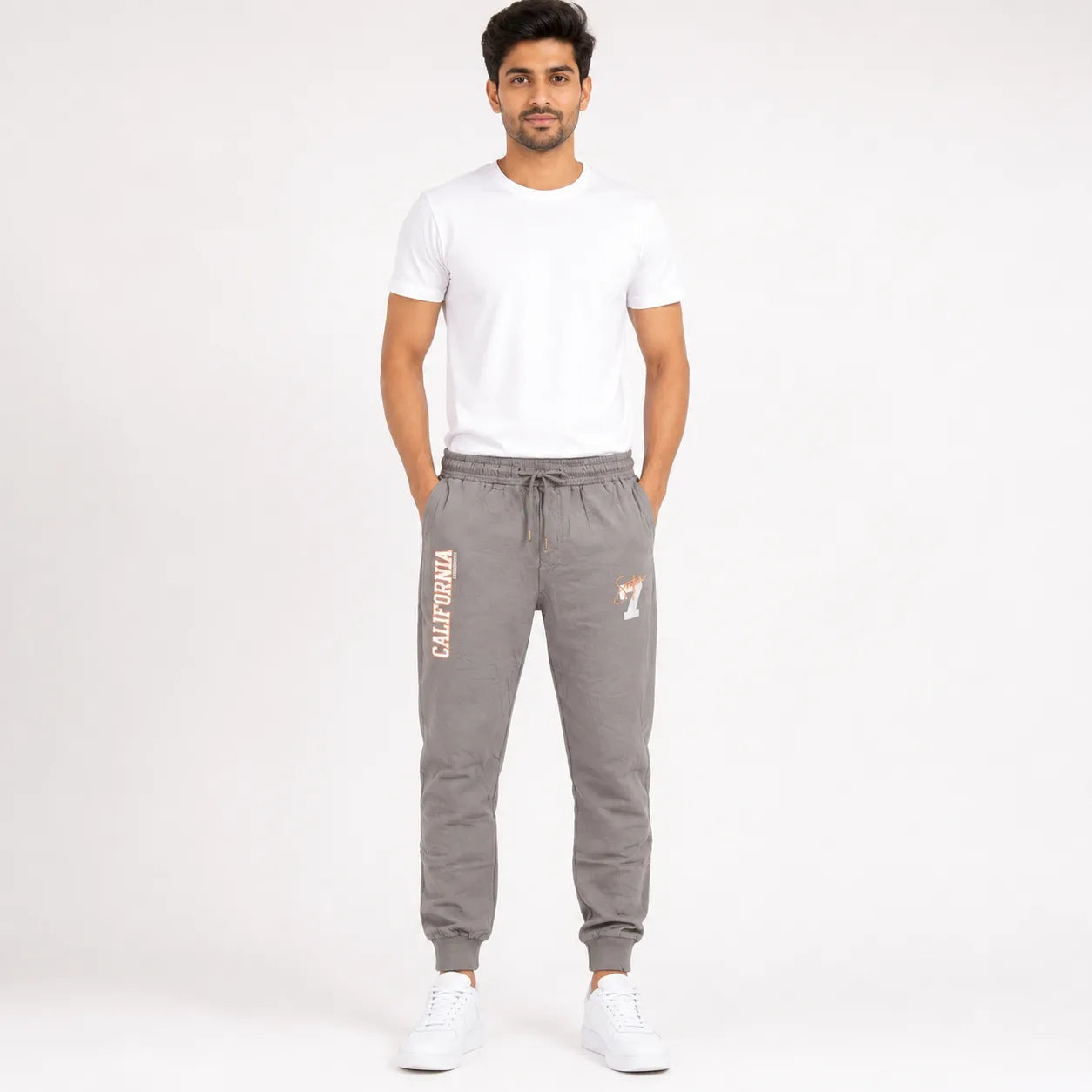 A man modeling gray sweatpants with 'CALIFORNIA' printed vertically on the left leg and a small logo on the right leg, paired with a white t-shirt and white sneakers against a plain white studio background.