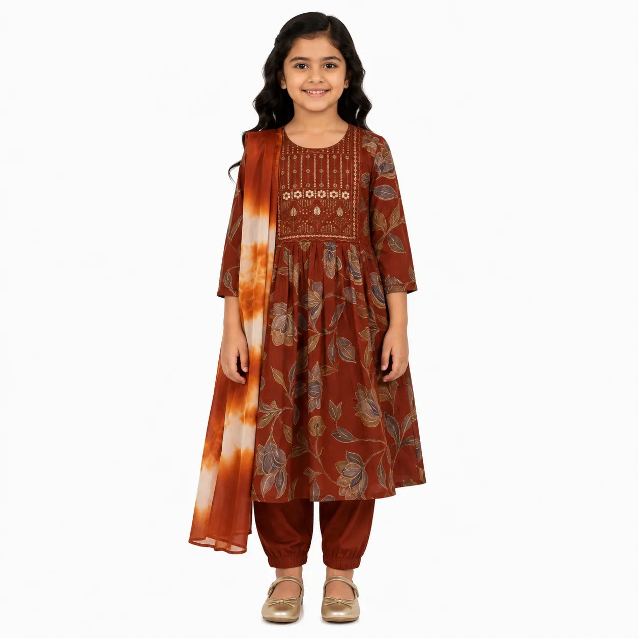 A young girl modeling a rust-brown floral printed kurta set with matching pants and a tie-dye dupatta, standing against a plain white background.