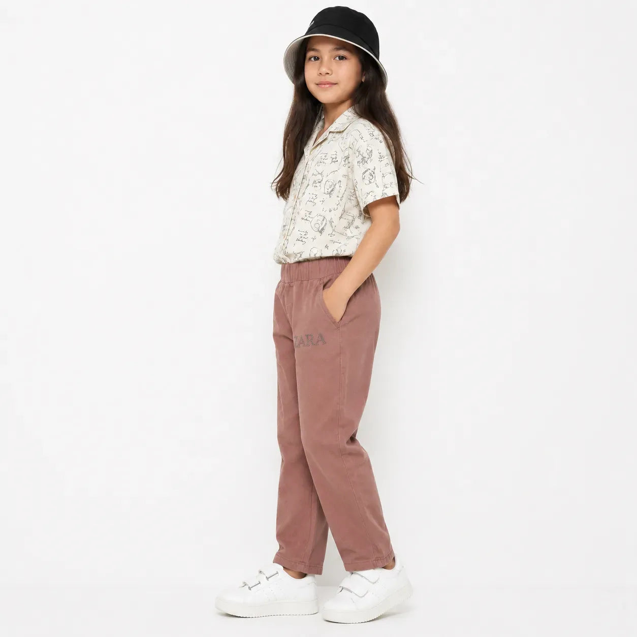 A young girl modeling a casual outfit consisting of dusty rose corduroy pants, a cream short-sleeve button-up shirt with black patterns, a black bucket hat, and white sneakers, standing against a plain white studio background.