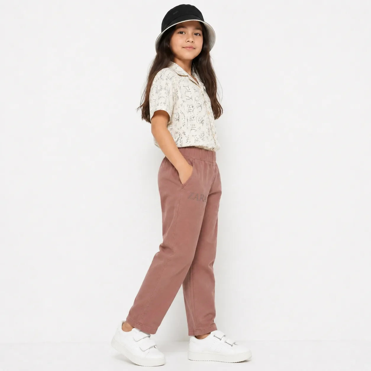 A young girl wearing dusty rose corduroy trousers with a subtle 'ZARA' logo on the thigh, paired with a white patterned short-sleeve shirt, white sneakers, and a black bucket hat, standing against a plain white studio background.