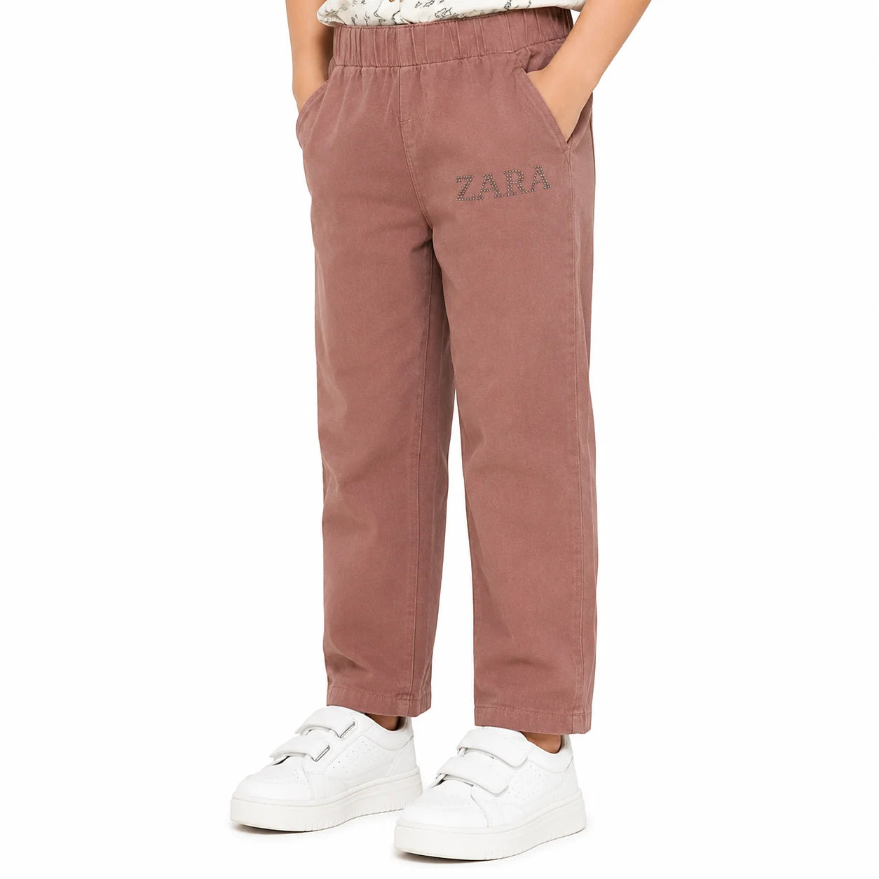 A child wearing dusty rose pants with a gray embroidered 'ZARA' logo on the left thigh, paired with white sneakers, shown from the waist down on a white background.