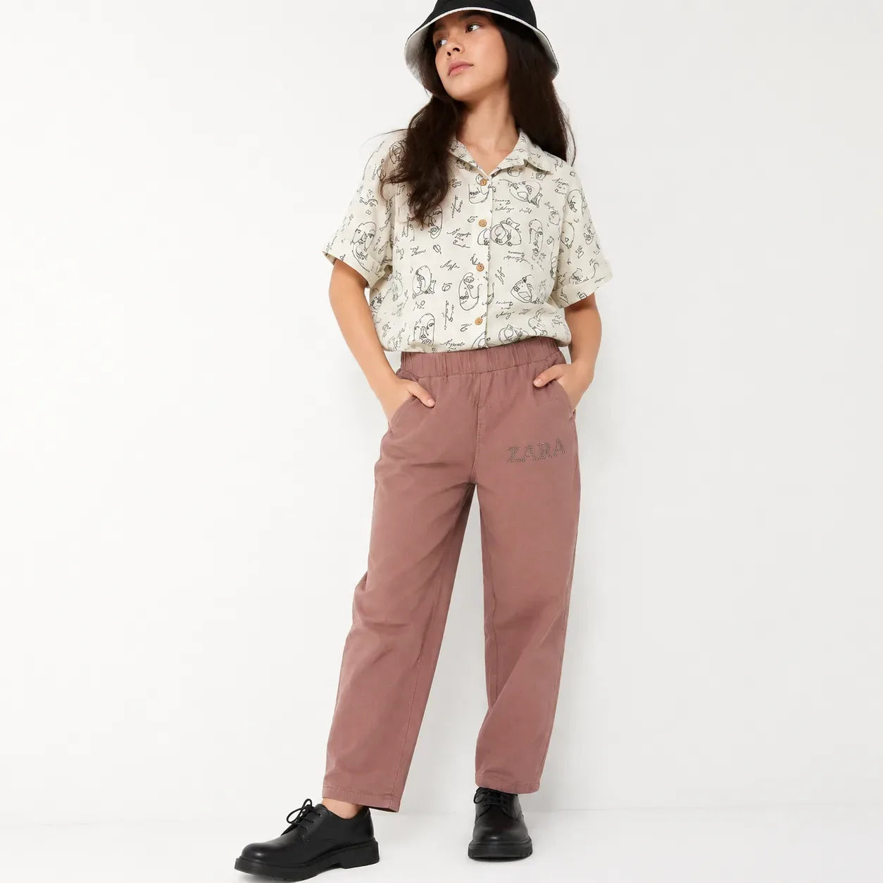 A young girl modeling a short-sleeve button-up shirt with a black line art animal print on an off-white background, paired with dusty rose wide-leg trousers featuring a subtle 'ZARA' logo on the left thigh, a black bucket hat, and black shoes, set against a plain white studio background.