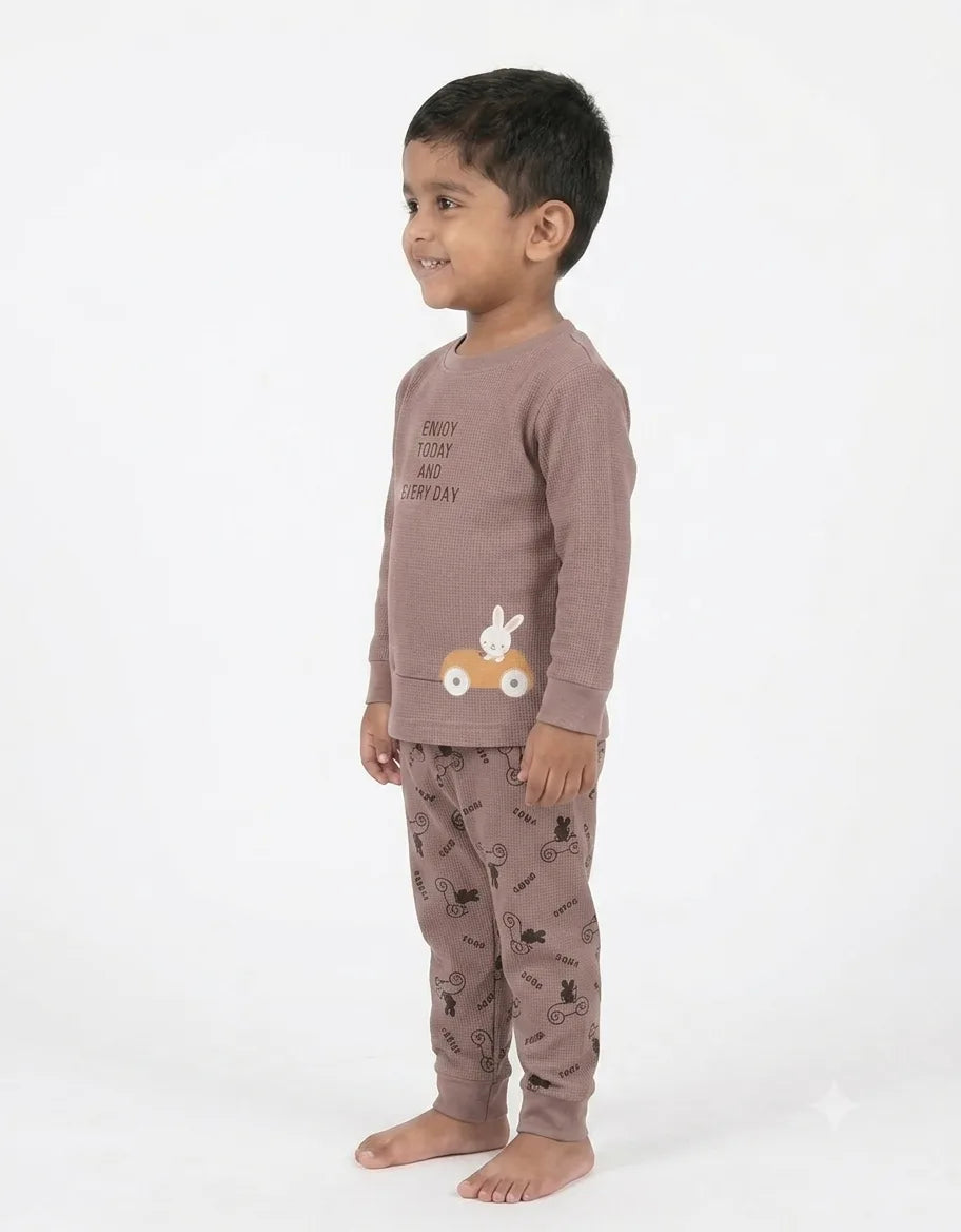 A toddler boy wearing a two-piece pajama set in a dusty mauve-brown color. The set includes a long-sleeve top with a bunny and car graphic, and matching patterned pants.