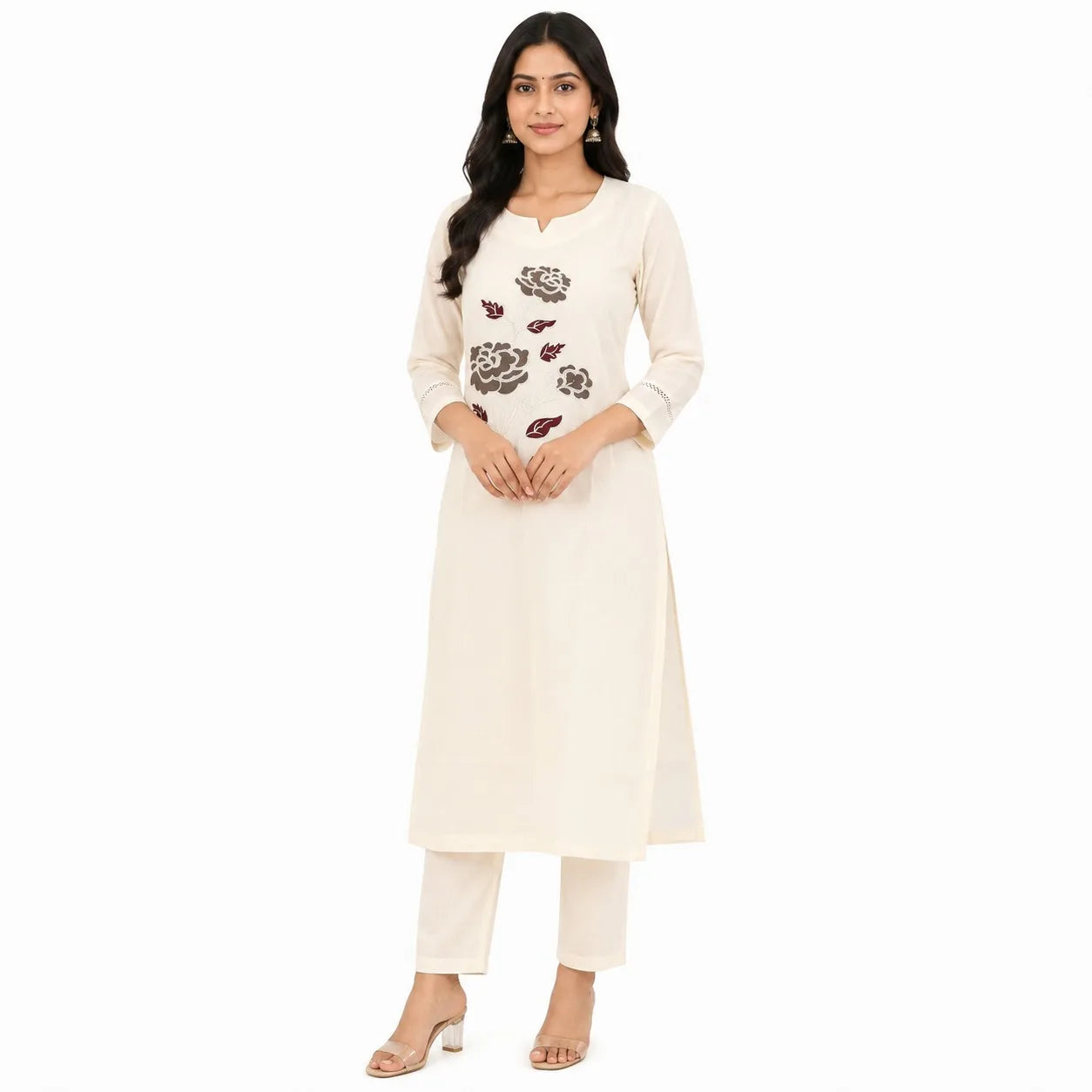 A woman modeling a cream-colored three-piece Indian ethnic suit, featuring a long kurta with floral embroidery on the chest, matching trousers, and heeled sandals, set against a plain white background.