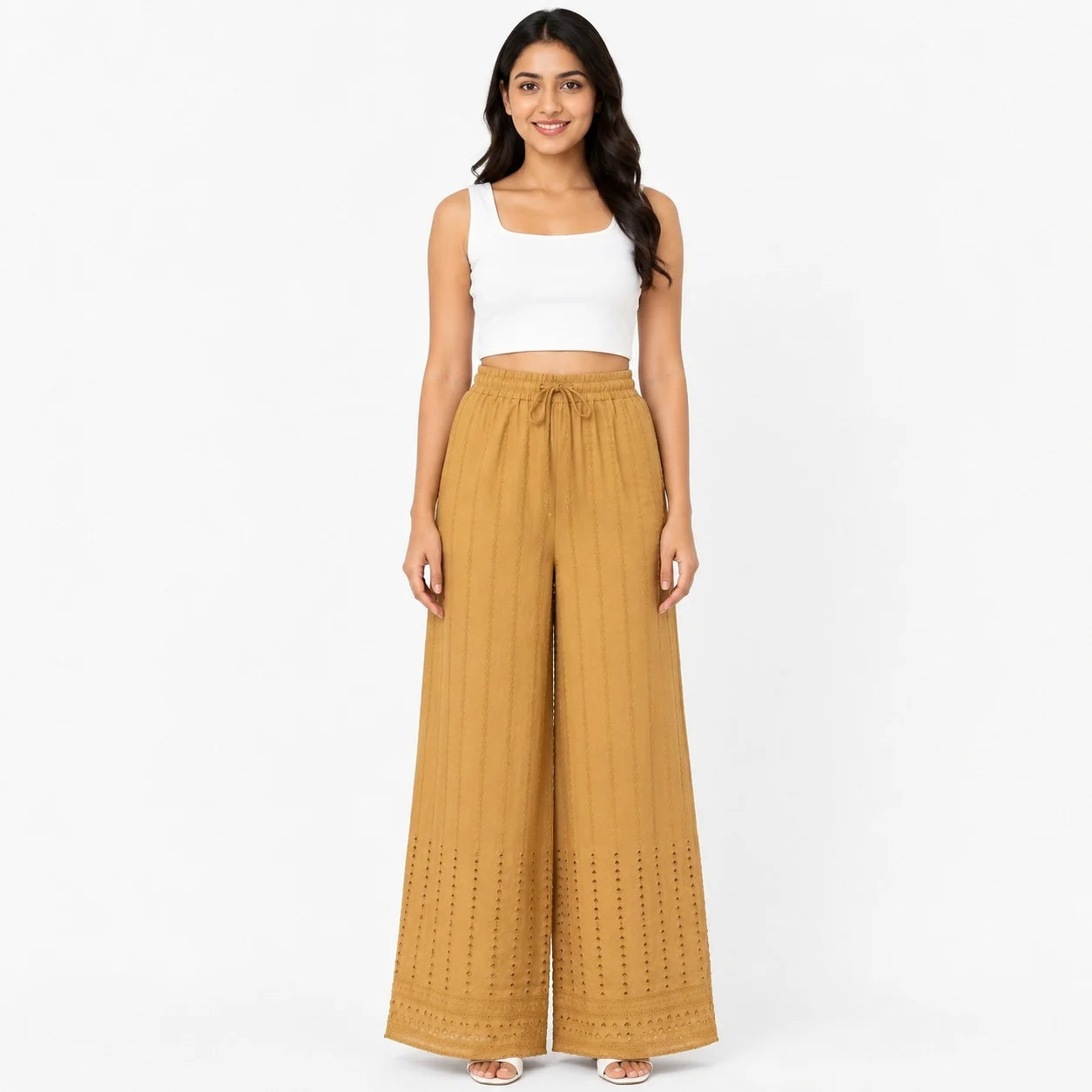 A woman modeling a pair of wide-leg, ankle-length pants in a warm mustard yellow color with vertical pleats and eyelet embroidery near the hems, paired with a white crop top against a plain white background.