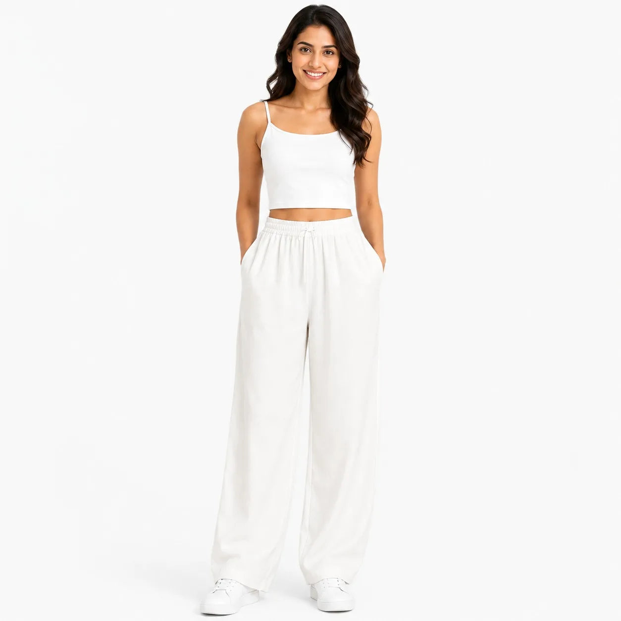 A woman modeling a white two-piece outfit consisting of a cropped camisole top and matching wide-leg pants, paired with white sneakers, against a solid white background.