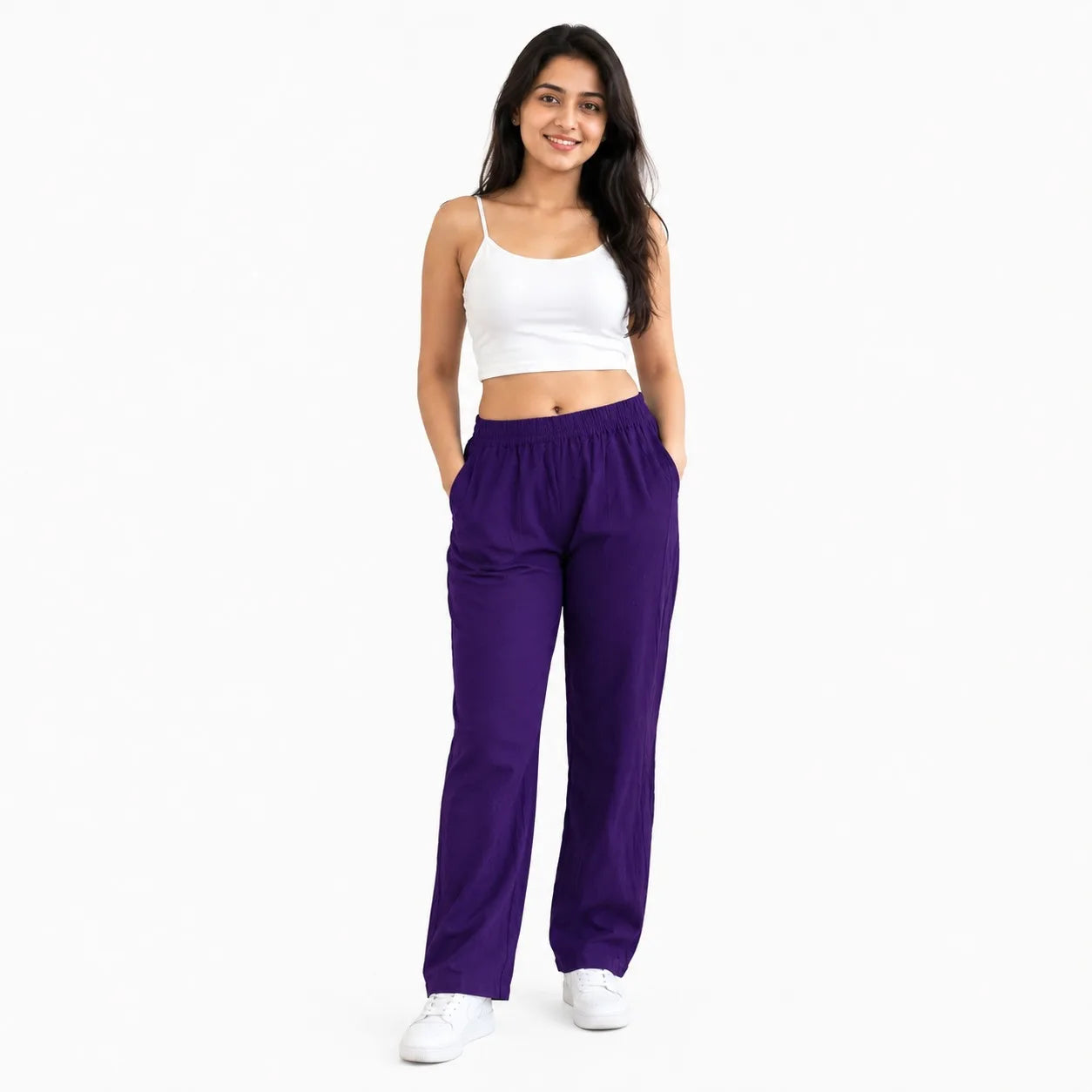 A woman modeling a pair of loose-fitting purple sweatpants with a white crop top and white sneakers, standing against a plain white background.