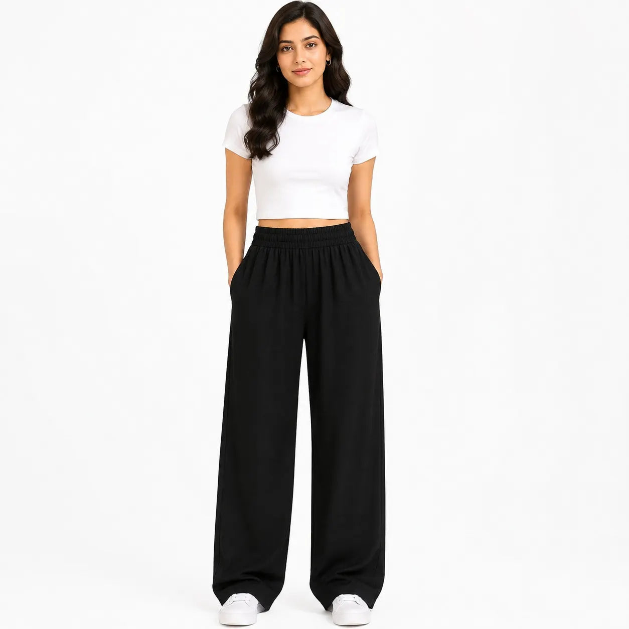 A woman modeling black wide-leg sweatpants with a matching black elastic waistband, paired with a white crop top and white sneakers, against a plain white studio background.
