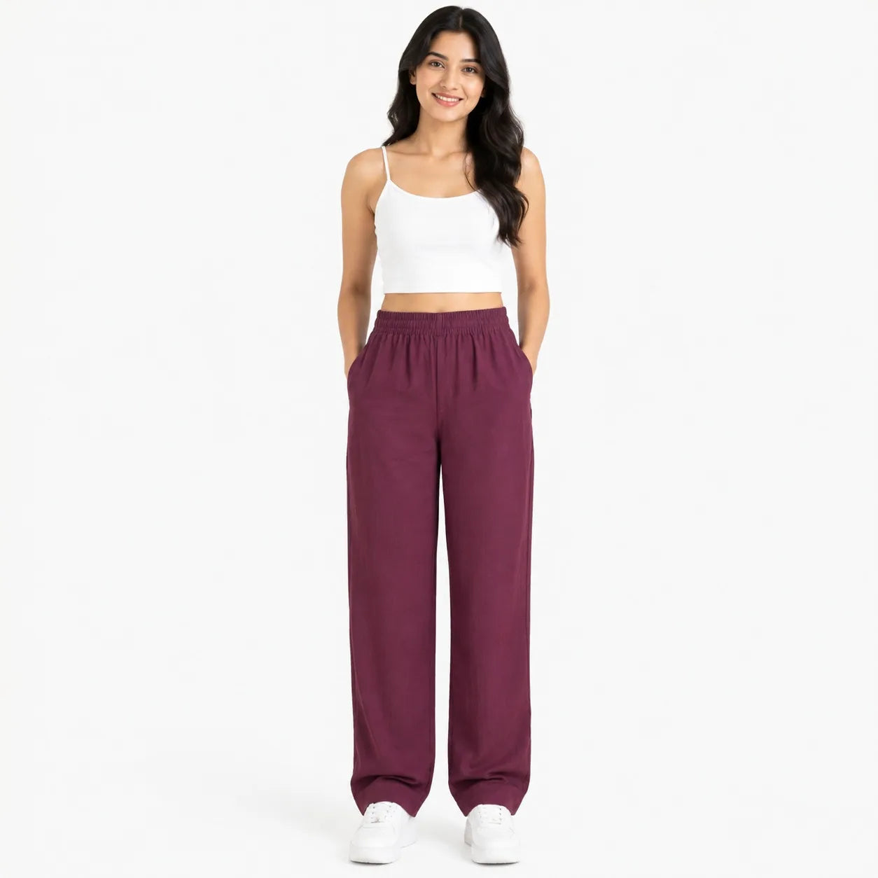 A woman modeling a pair of wide-leg, elastic-waist pants in a deep plum or burgundy shade, paired with a white spaghetti-strap crop top and white sneakers, against a plain white studio background.