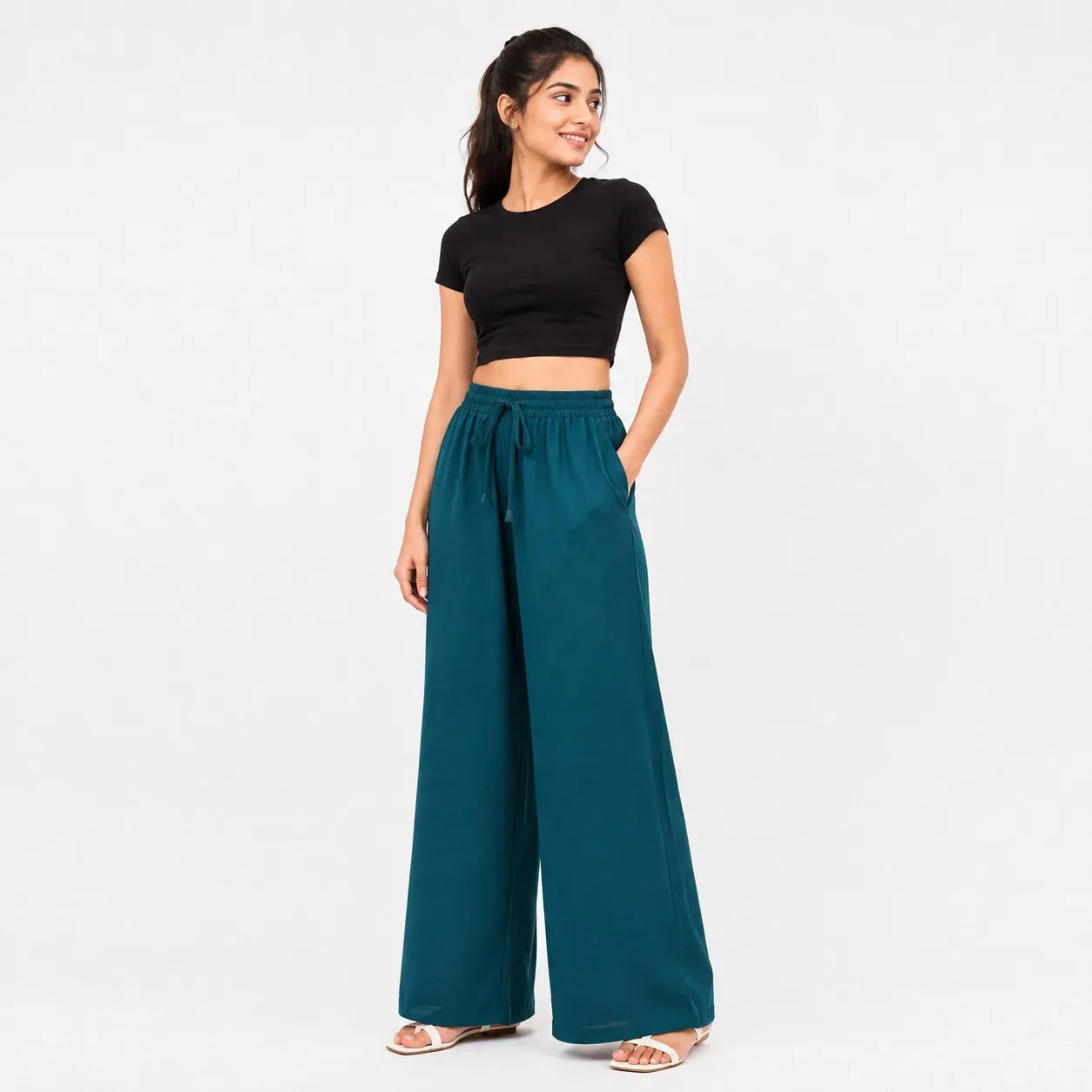 A woman modeling a black short-sleeve crop top and teal wide-leg drawstring pants against a plain white studio background.