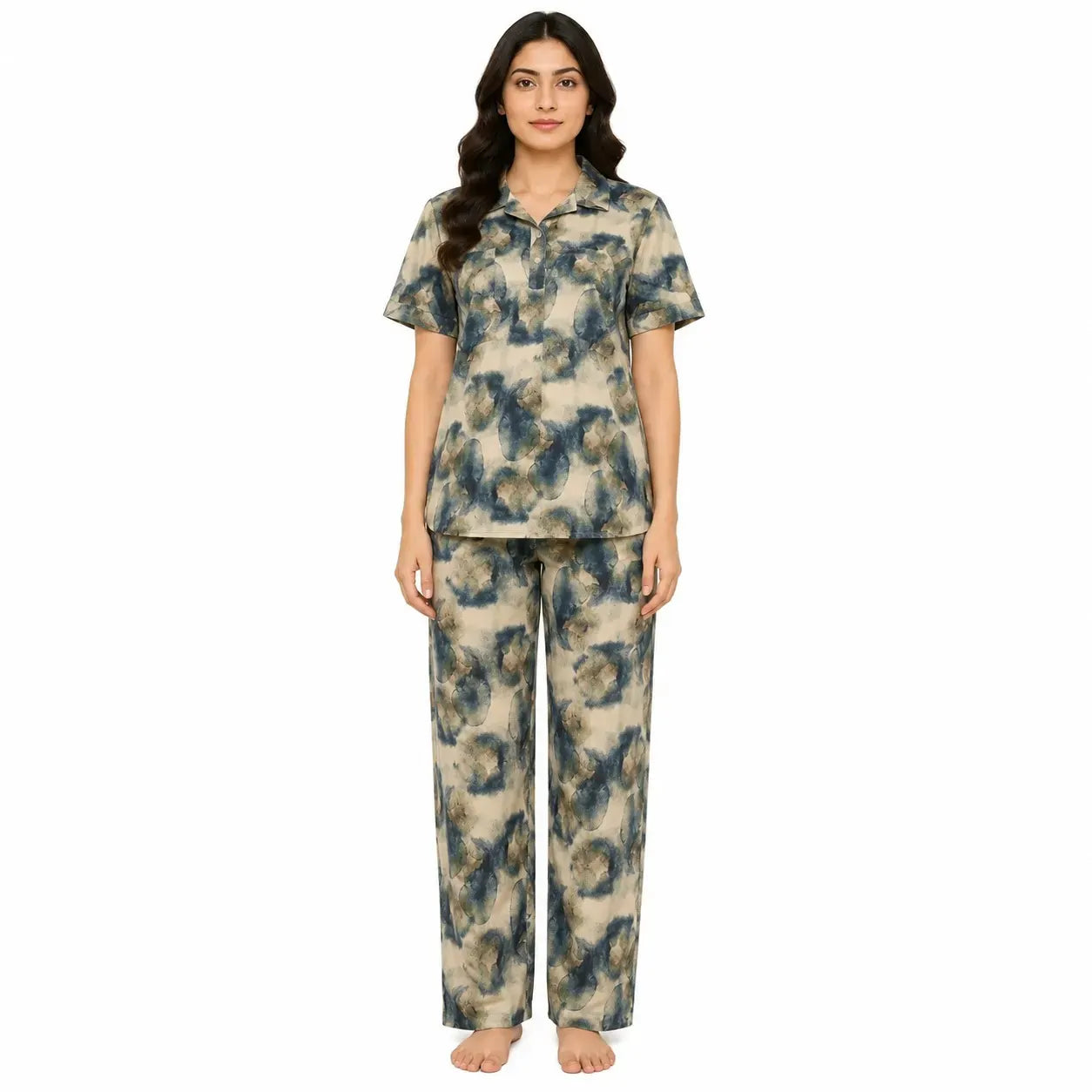 A woman modeling a two-piece pajama set with a short-sleeve collared top and matching pants, featuring an abstract blue and gray pattern on a beige background, against a plain white studio backdrop.