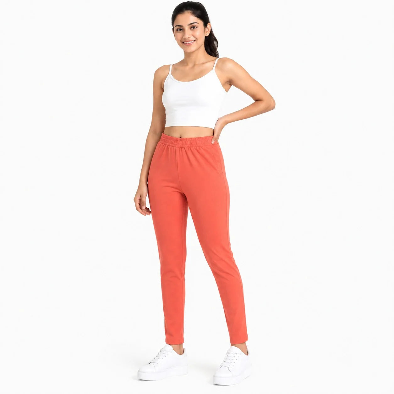 A woman modeling a pair of high-waisted, slim-fit sweatpants in a vibrant coral orange color, paired with a white crop top and white sneakers, against a plain white background.