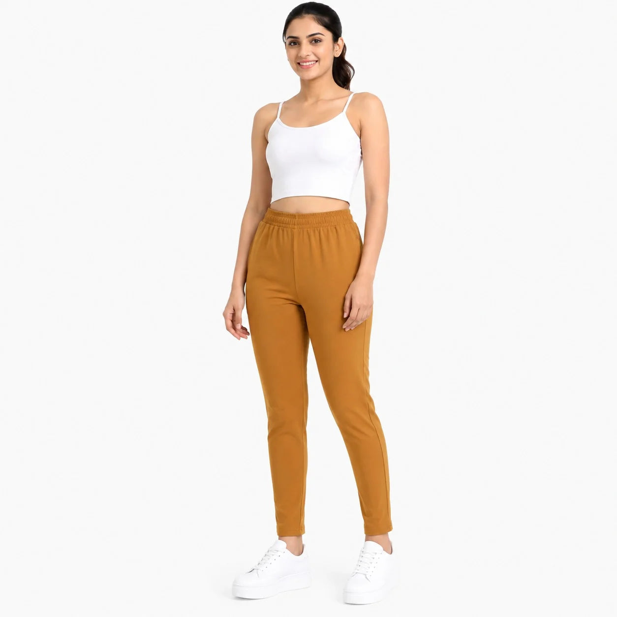 A woman modeling a two-piece activewear set consisting of a white ribbed crop tank top and high-waisted, tapered jogger pants in a warm camel-brown color, paired with white platform sneakers, against a solid white background.