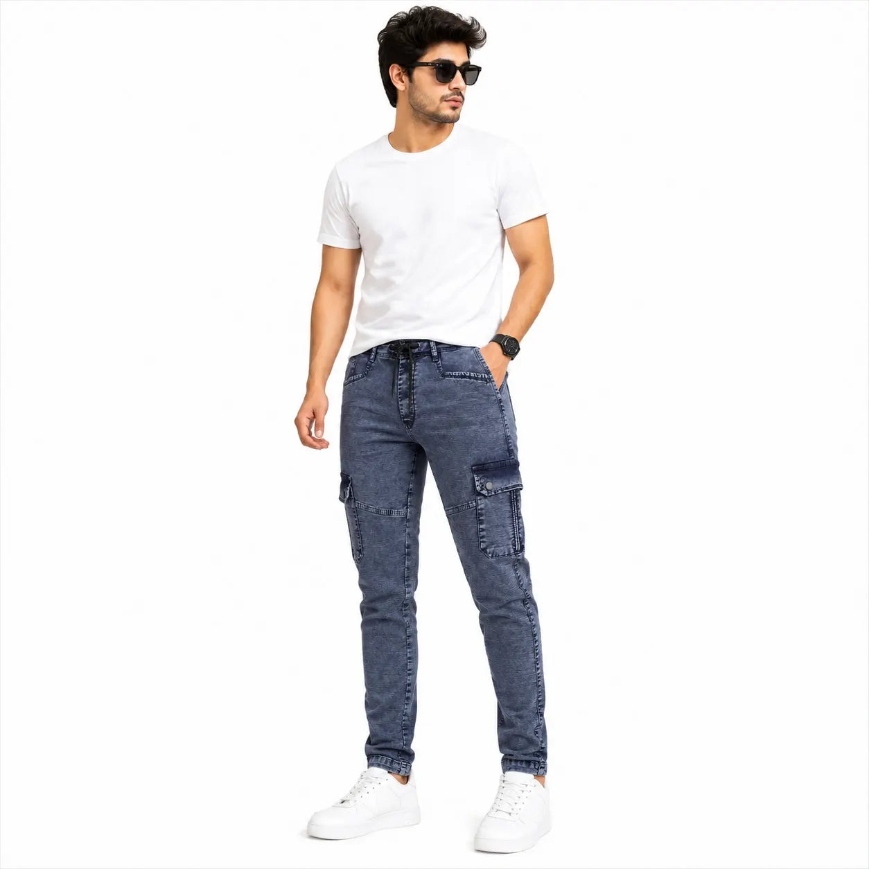 A man modeling a pair of dark blue slim-fit cargo jeans, styled with a white t-shirt, white sneakers, and sunglasses, set against a plain white background.
