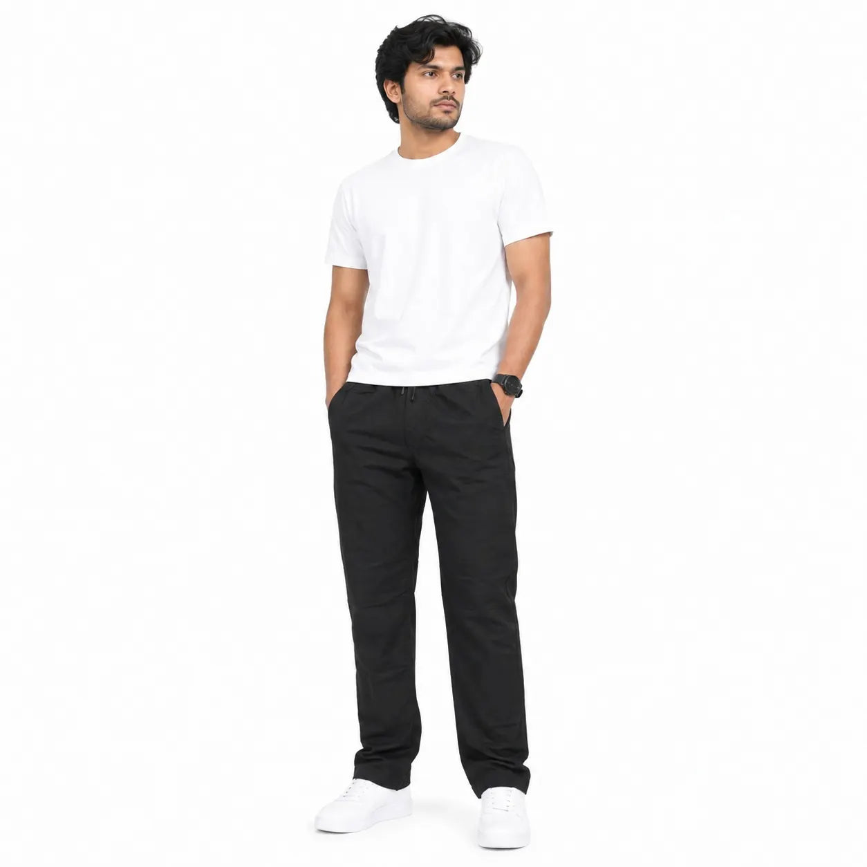 A man wearing a plain white crew-neck t-shirt, black trousers, and white sneakers, standing against a solid white background.