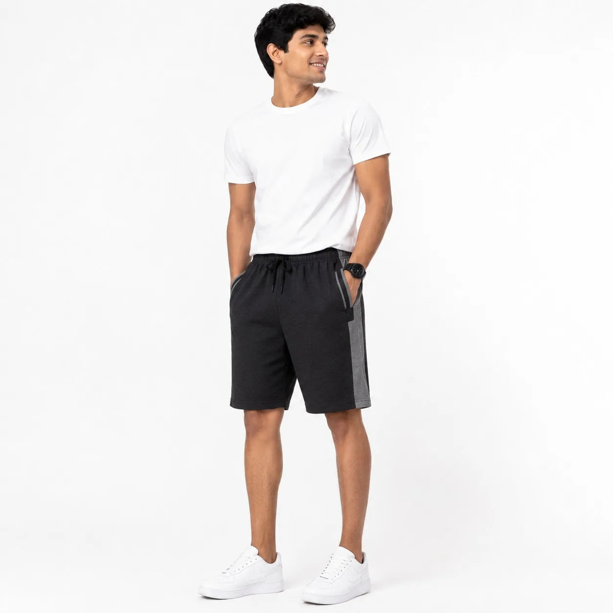 A man modeling black athletic shorts with gray side panels, paired with a white t-shirt and white sneakers, standing against a plain white studio background.