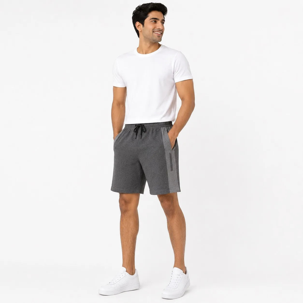 A man modeling dark gray athletic shorts with a side pocket, paired with a white t-shirt and white sneakers, standing against a plain white studio background.