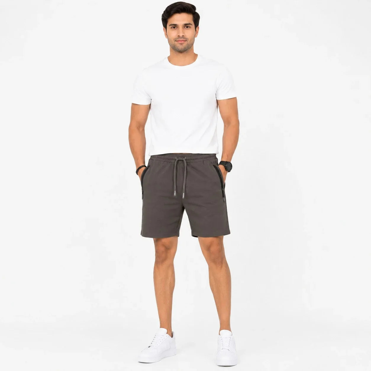 A man modeling dark gray drawstring shorts, paired with a white t-shirt and white sneakers, standing against a plain white studio background.