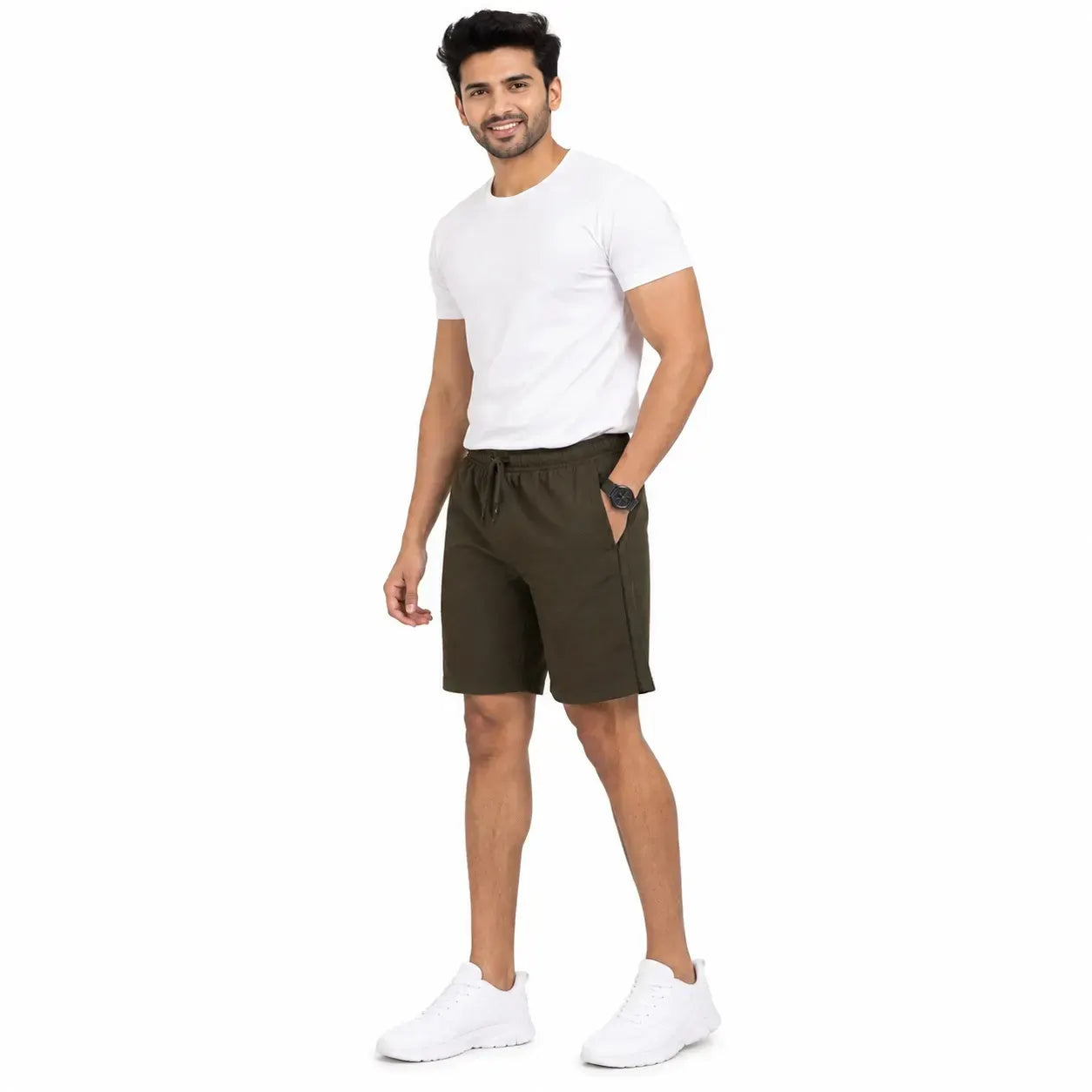 A man modeling dark olive green athletic shorts, paired with a white t-shirt and white sneakers, standing against a solid white background.