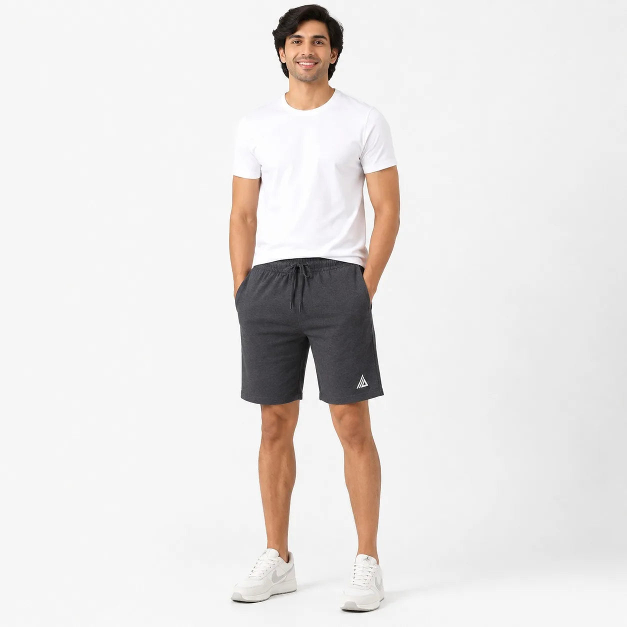 A man modeling dark gray athletic shorts with a white drawstring, paired with a white t-shirt and white sneakers, standing against a plain white studio background.