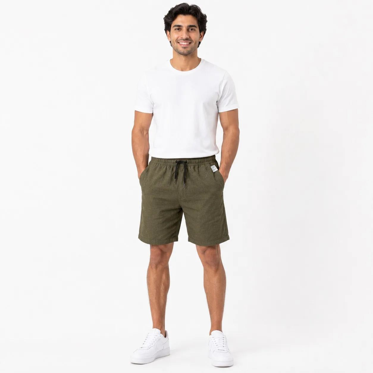 A man modeling olive green linen drawstring shorts, paired with a white t-shirt and white sneakers, standing against a plain white studio background.