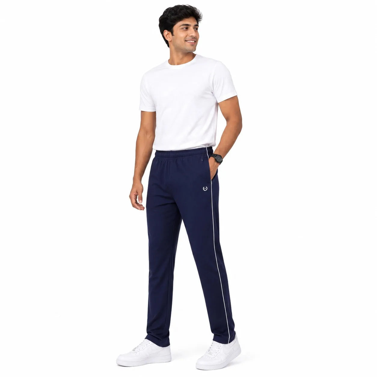 A man modeling navy blue track pants with a white side stripe and a small white embroidered logo, paired with a white t-shirt and white sneakers, on a solid white background.