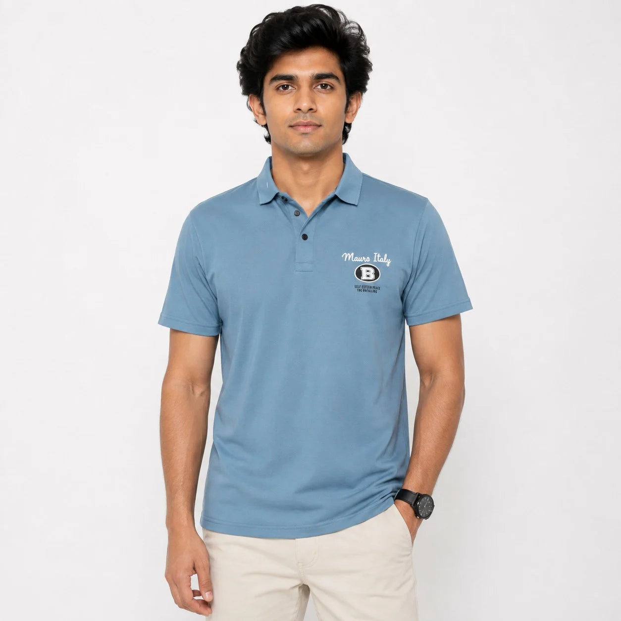 A man wearing a light blue short-sleeve polo shirt with a black and white logo on the left chest, paired with beige trousers, standing against a plain white background.