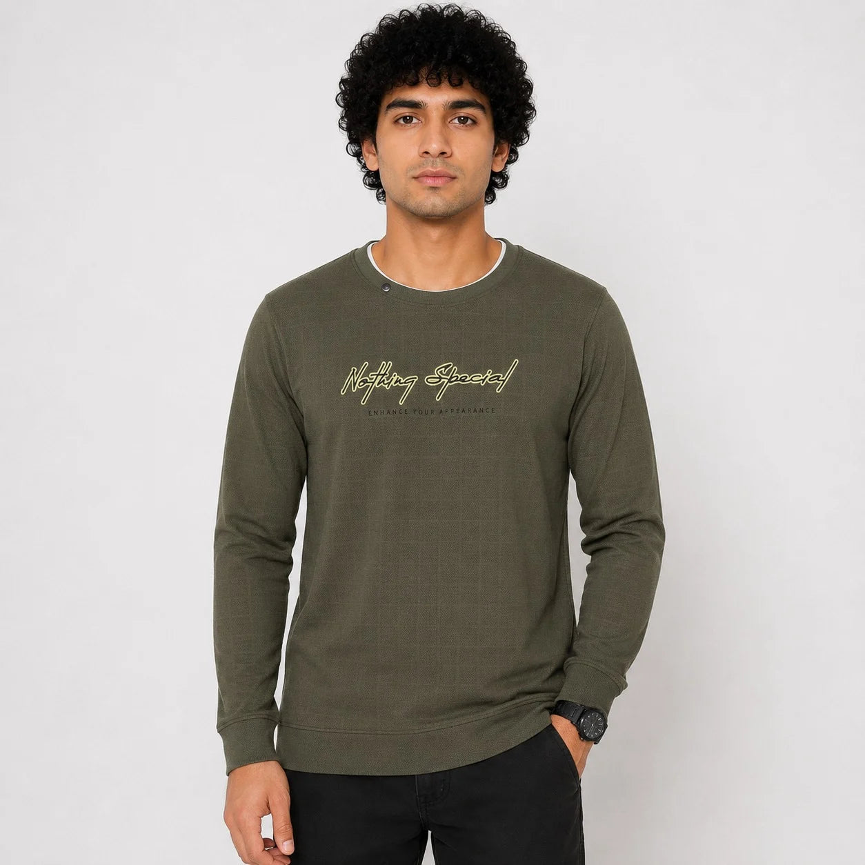 A man wearing an olive green long-sleeve crewneck sweatshirt with 'Nothing Wrong' printed in gold script on the chest, paired with black pants, against a light gray studio background.