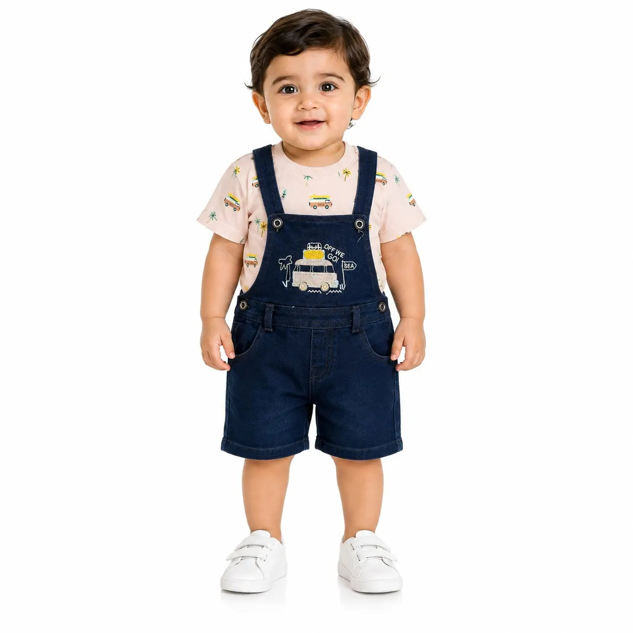 A toddler standing against a white background, wearing a light pink short-sleeve t-shirt with a small yellow and green train pattern, dark blue denim overall shorts, and white sneakers.