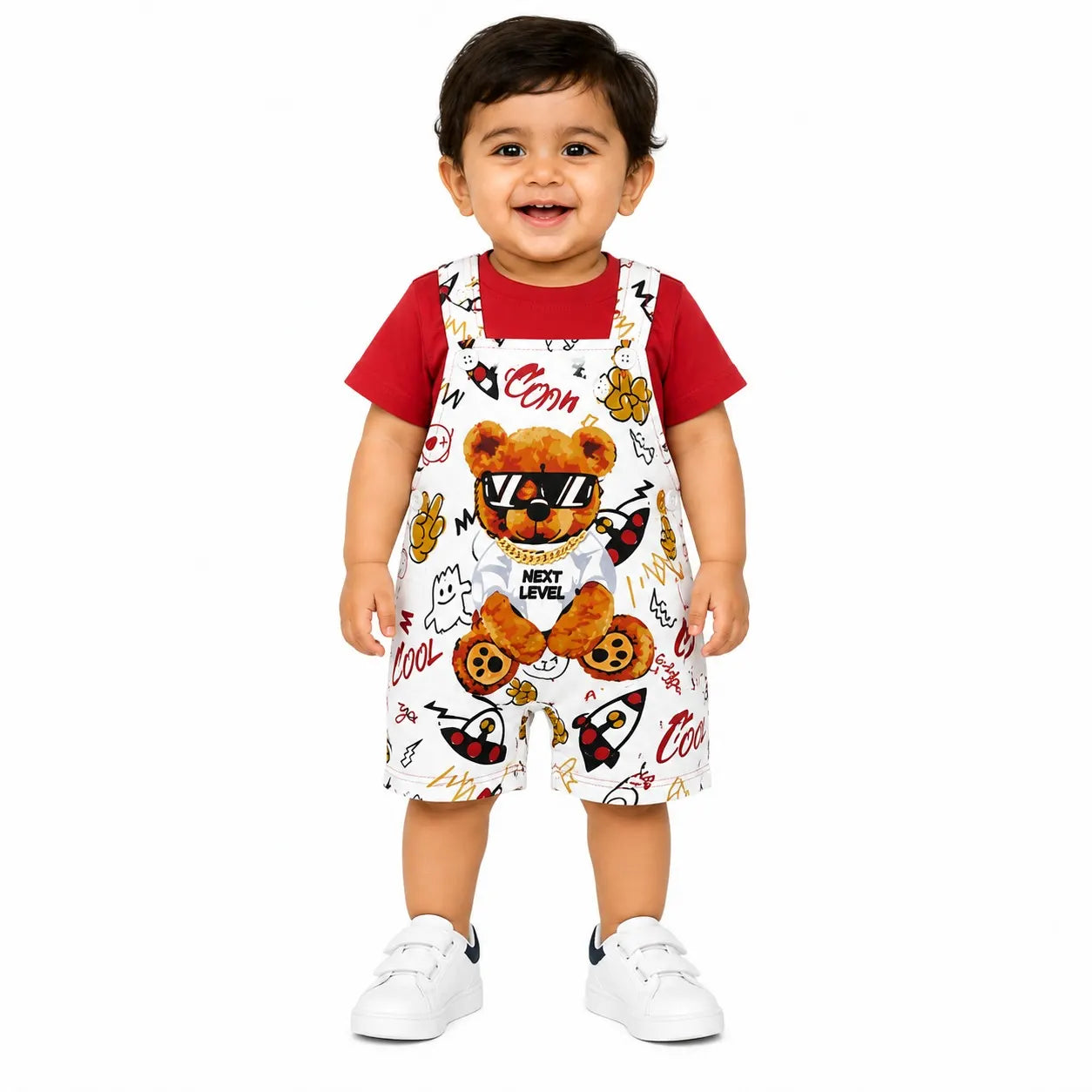 A toddler standing against a white background wearing a red short-sleeve shirt and white bib-style overalls with a teddy bear and cartoon print, paired with white sneakers.
