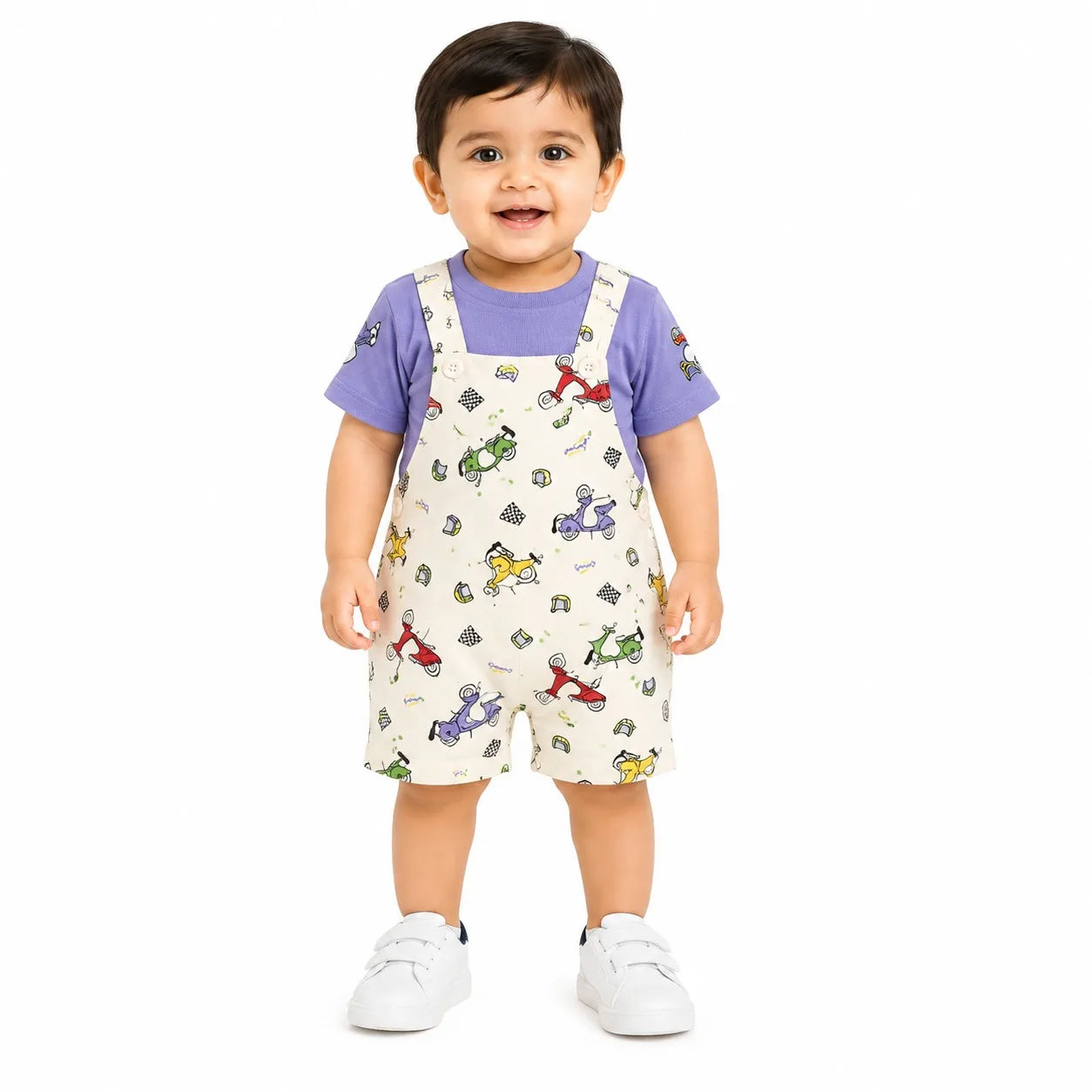 A toddler standing against a white background wearing a purple short-sleeve t-shirt and cream-colored overalls covered in a colorful toy pattern, paired with white sneakers.