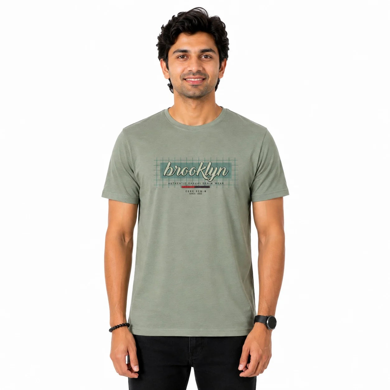 A man wearing a muted sage green t-shirt with a retro-style 'Brooklyn' graphic on the chest, paired with black pants, standing against a plain white background.