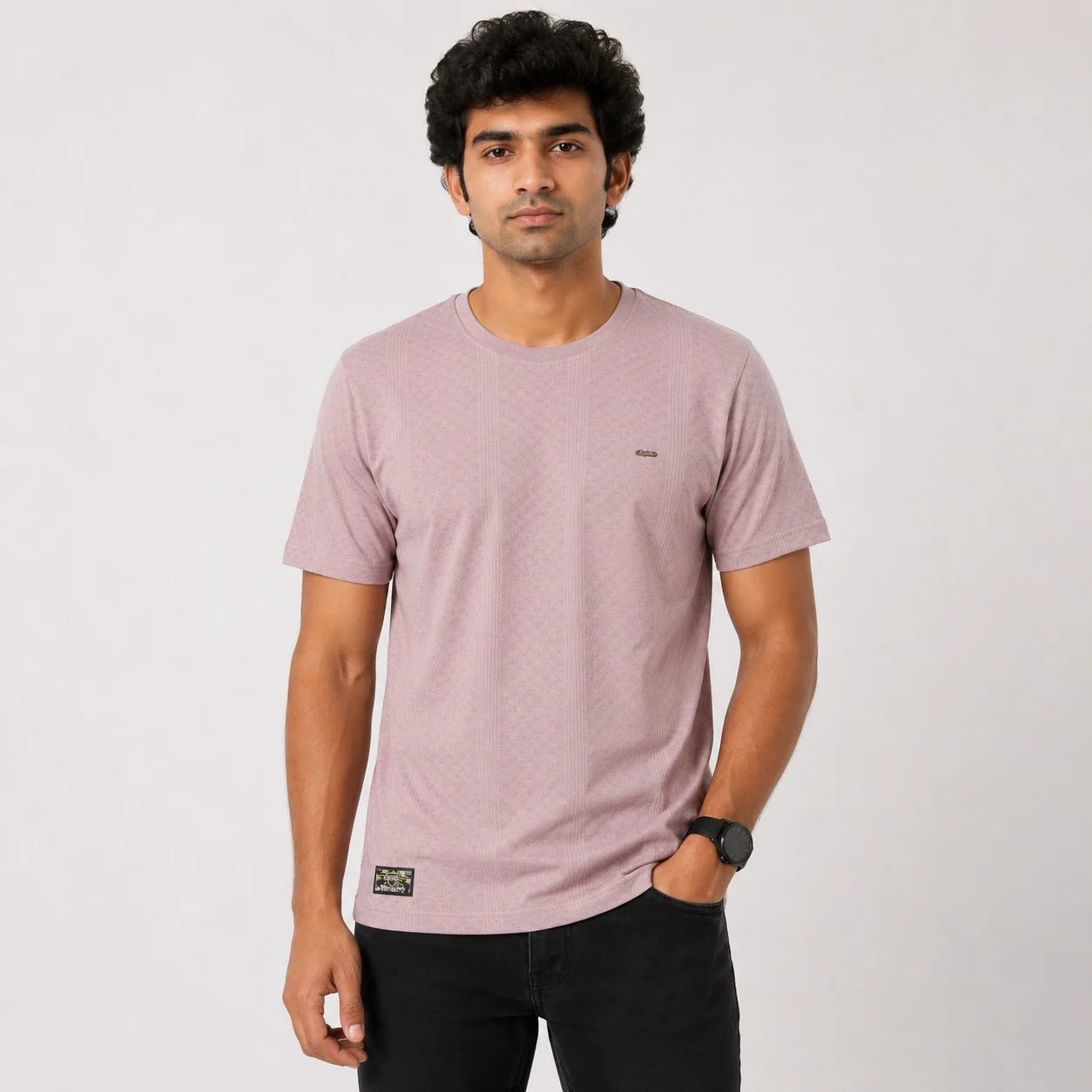 A man wearing a dusty pink short-sleeve crewneck t-shirt with a small embroidered logo on the left chest and a brand patch near the bottom hem, paired with black pants, against a plain light gray background.