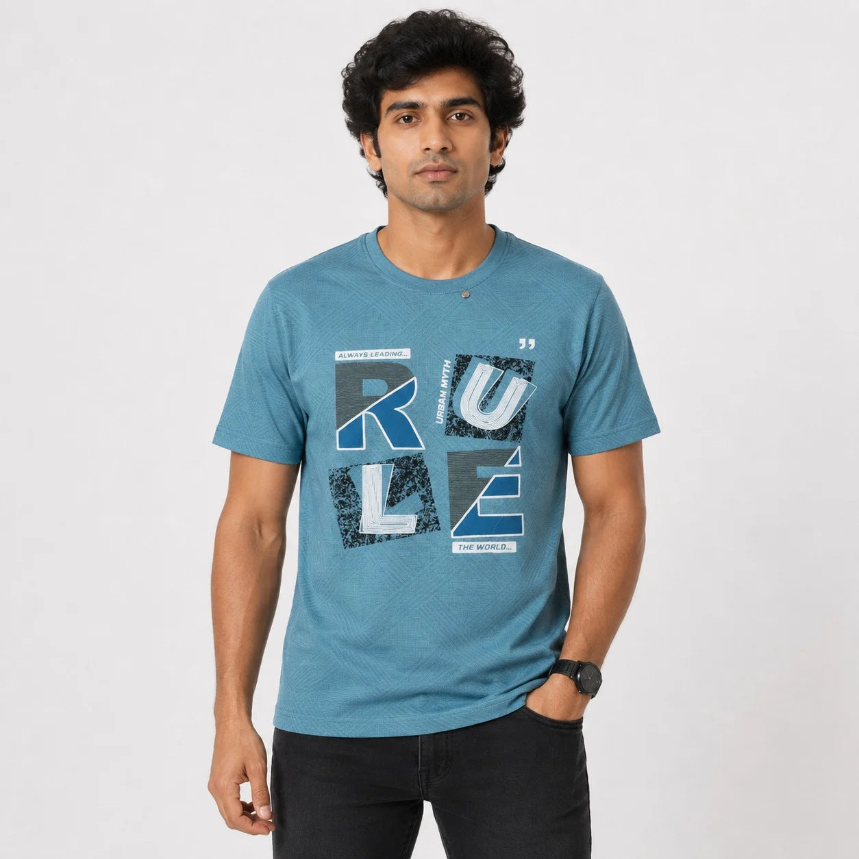 A man wearing a heathered blue crewneck t-shirt with a graphic print of the word 'RULE' in distressed gray and white blocks, paired with black pants, standing against a light gray studio background.